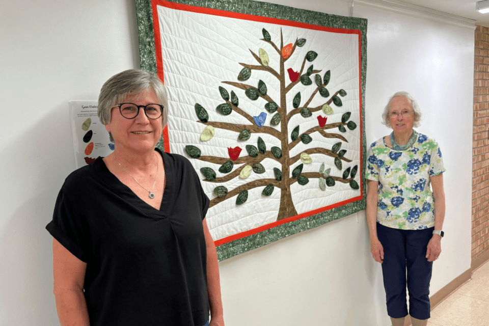 Two women standing next to a quilt wall hanging